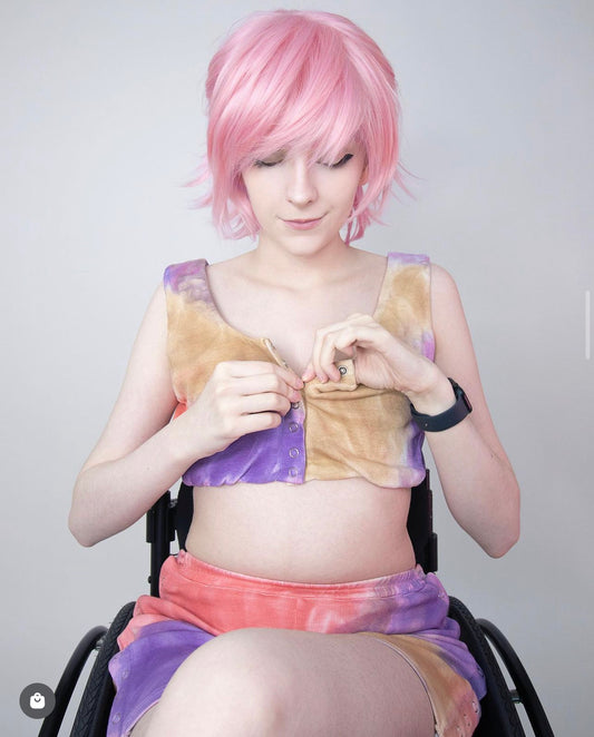 Women in wheelchair with pink hair wearing colorful adaptive top and shorts